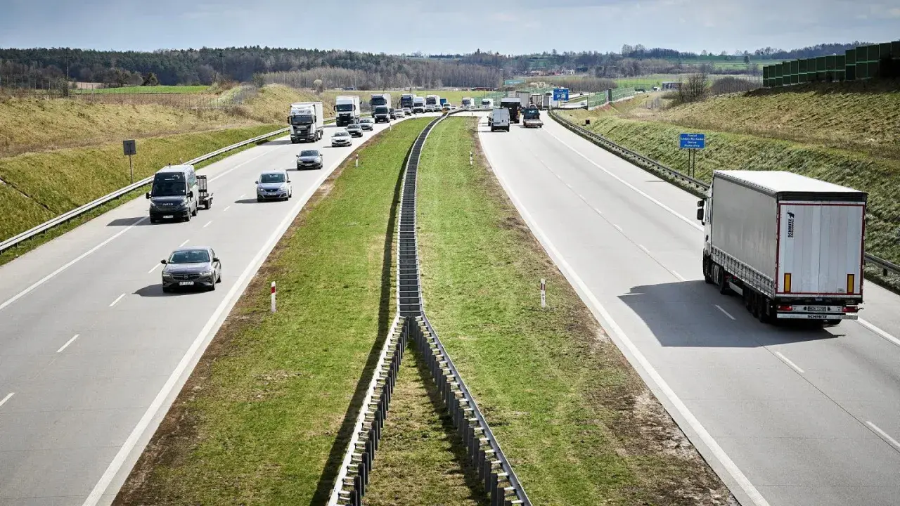 Ciężarówki na autostradzie pędzą z dużą prędkością, pokonując kolejne kilometry. W tle widać inne pojazdy i krajobraz.