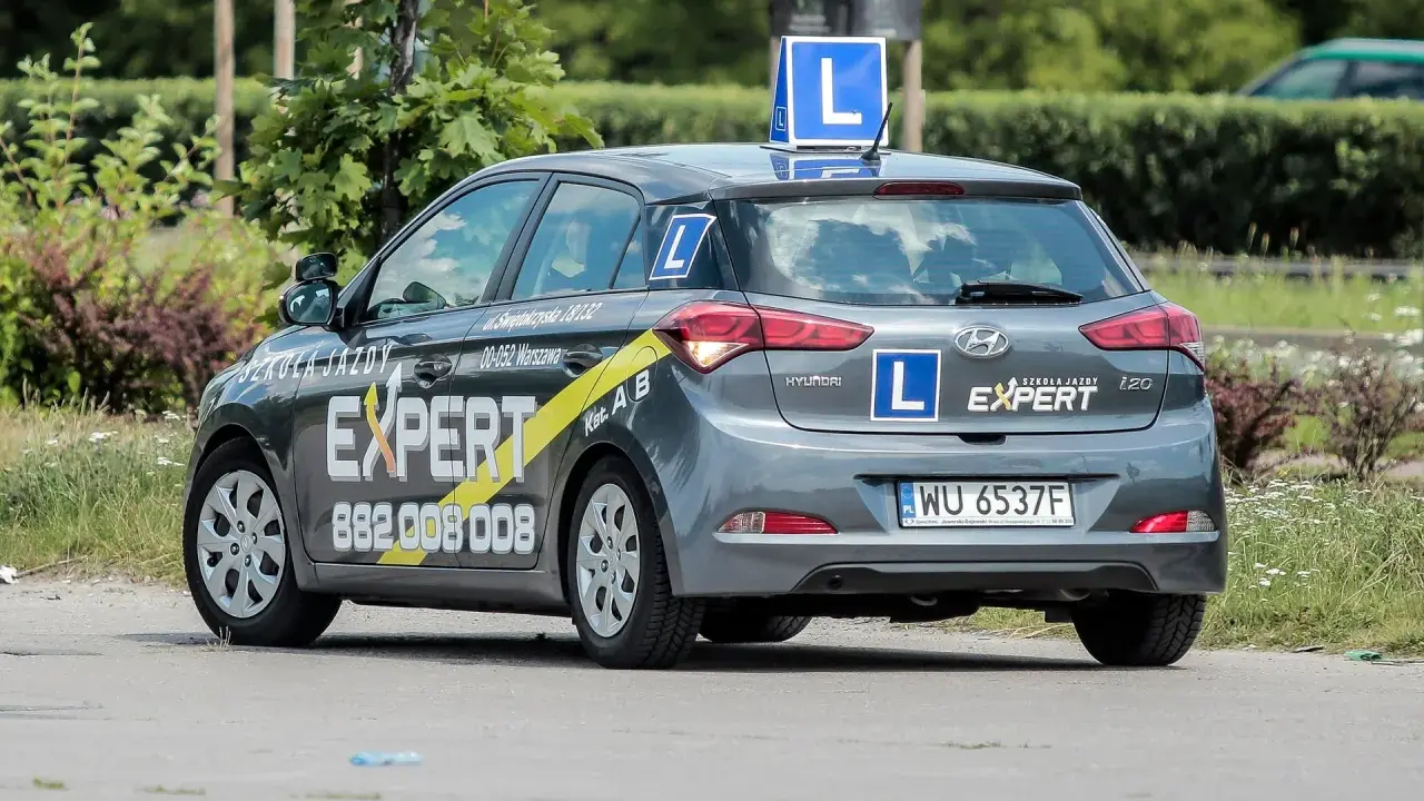 Szara Skoda i20 z oznaczeniami "Szkoła Jazdy EXPERT" i literą "L" na dachu i bokach. To auto do nauki jazdy, idealne dla przyszłego kierowcy.