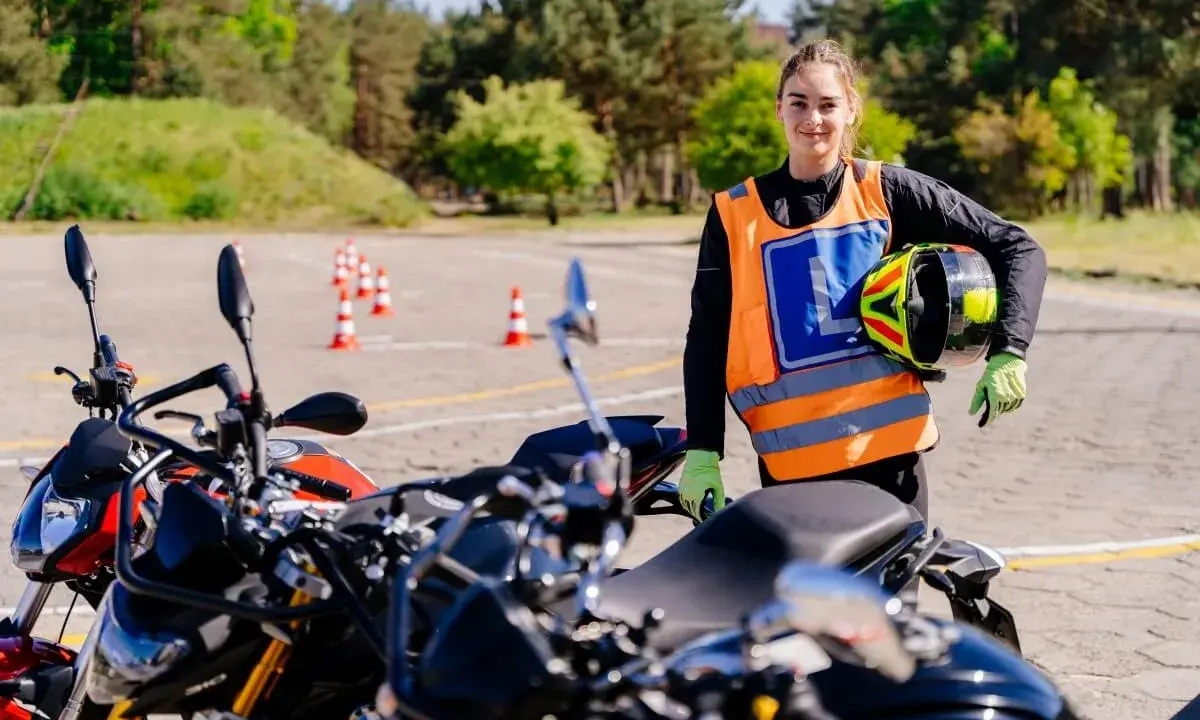 Młoda kobieta w kamizelce z literą "L" i kaskiem w ręku, gotowa do jazdy na motocyklu na placu manewrowym. To jej pierwsze kroki na drodze, a wkrótce będzie mogła jeździć na A1.