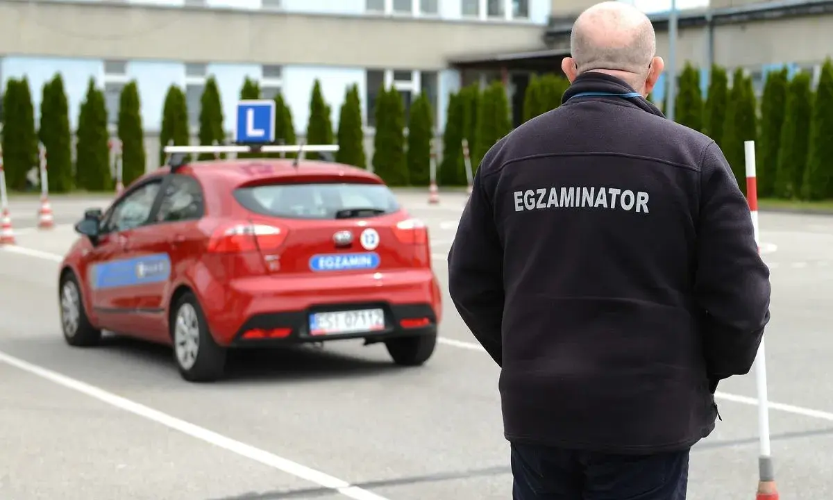 Egzaminator w czarnej kurtce z napisem "EGZAMINATOR" obserwuje czerwony samochód z literą "L" na dachu. Jak odwołać egzamin na prawo jazdy infocar?
