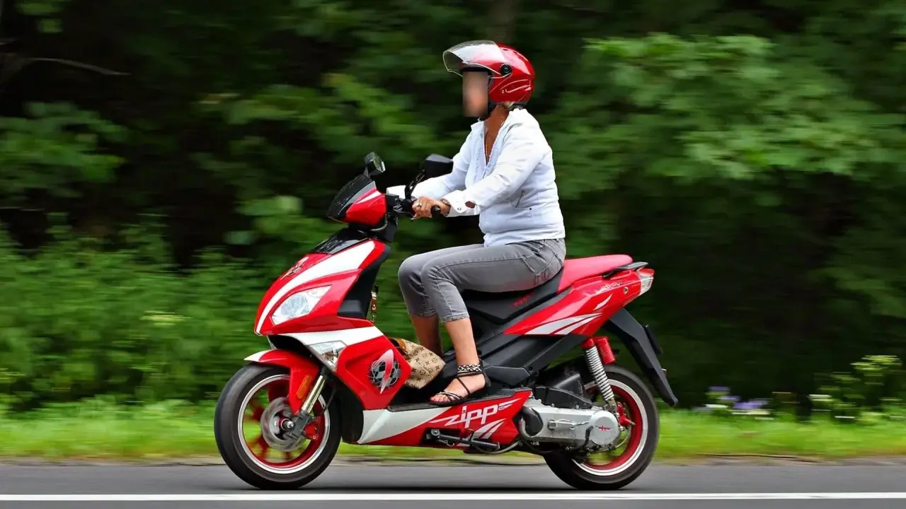 Kobieta w kasku jedzie czerwonym skuterem po autostradzie, otoczona zielenią.