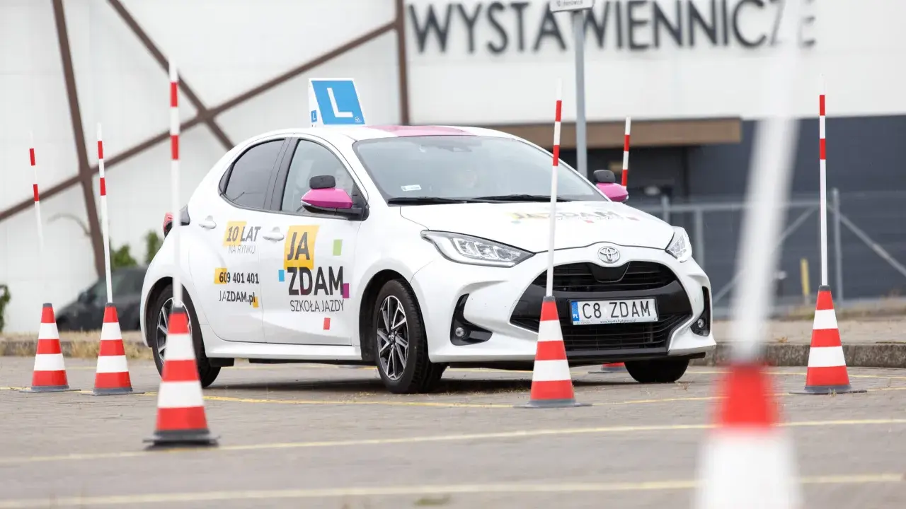 Biały samochód szkoleniowy z literą "L" na dachu i pachołkami na placu manewrowym. To miejsce, gdzie nauczysz się, jak wypełnić pkk.