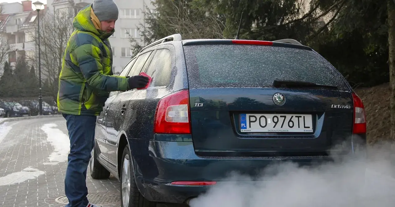 Mężczyzna w zimowej kurtce czyści zamarzniętą szybę auta. Dym z rury wydechowej sugeruje, że to diesel i właśnie próbuje się dowiedzieć, jak odpalić diesla w mroźny dzień.