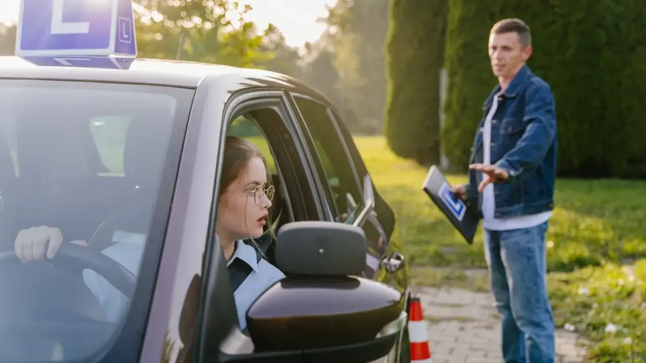 Młoda kobieta w okularach za kierownicą, z literą "L" na dachu, uczy się jeździć. Instruktor z tablicą daje wskazówki. To jej kontynuacja prawa jazdy po latach.