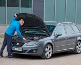 Mężczyzna w niebieskiej kurtce sprawdza silnik szarego kombi. To ważny moment przed kupnem samochodu, by upewnić się, że wszystko działa.