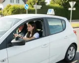 Młoda dziewczyna uczy się jeździć, a obok siedzi instruktor. Znak "L" na dachu sugeruje, że to nauka jazdy. Od kiedy można robić prawo jazdy?