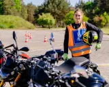 Młoda kobieta w kamizelce z literą "L" i kaskiem w ręku, gotowa do jazdy na motocyklu na placu manewrowym. To jej pierwsze kroki na drodze, a wkrótce będzie mogła jeździć na A1.