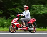 Kobieta w kasku jedzie czerwonym skuterem po autostradzie, otoczona zielenią.