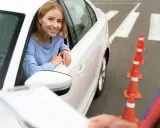 Uśmiechnięta kobieta w samochodzie czeka na rozpoczęcie egzaminu wewnętrznego na prawo jazdy. Przed nią stoją pomarańczowe pachołki.