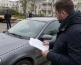 Mężczyzna czyta dokumenty przy samochodzie, zastanawiając się, jak odzyskać dowód rejestracyjny zatrzymany elektronicznie. W tle widać lawetę i policjantów.