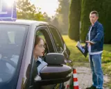 Młoda kobieta w okularach za kierownicą, z literą "L" na dachu, uczy się jeździć. Instruktor z tablicą daje wskazówki. To jej kontynuacja prawa jazdy po latach.