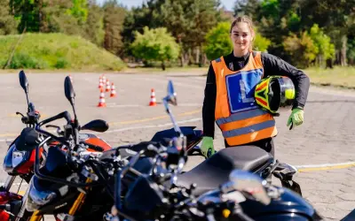 Młoda kobieta w kamizelce z literą "L" i kaskiem w ręku, gotowa do jazdy na motocyklu na placu manewrowym. To jej pierwsze kroki na drodze, a wkrótce będzie mogła jeździć na A1.