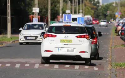 Samochody z literą "L" na dachu i tabliczką "EGZAMIN" czekają na egzamin na prawo jazdy.
