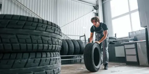 Mechanik w warsztacie przygotowuje opony zimowe przy 20 stopniach. Stosy opon czekają na wymianę.