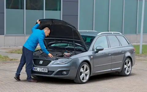 Mężczyzna w niebieskiej kurtce sprawdza silnik szarego kombi. To ważny moment przed kupnem samochodu, by upewnić się, że wszystko działa.