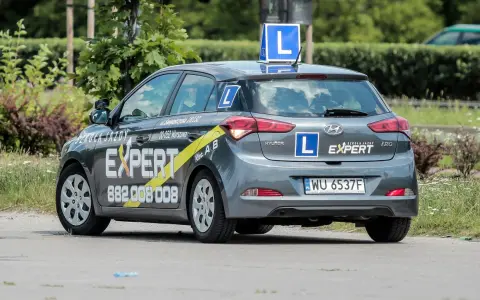 Szara Skoda i20 z oznaczeniami "Szkoła Jazdy EXPERT" i literą "L" na dachu i bokach. To auto do nauki jazdy, idealne dla przyszłego kierowcy.