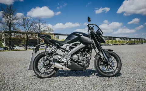 Czarny motocykl Yamaha MT-125, model kat a1, zaparkowany na żwirowym placu przed budynkiem.