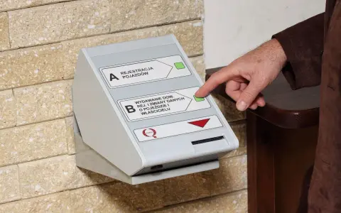 Dłoń naciska przycisk na automacie do wydawania dowodów rejestracyjnych, ułatwiając wymianę dowodu rejestracyjnego online.