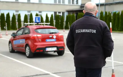 Egzaminator w czarnej kurtce z napisem "EGZAMINATOR" obserwuje czerwony samochód z literą "L" na dachu. Jak odwołać egzamin na prawo jazdy infocar?