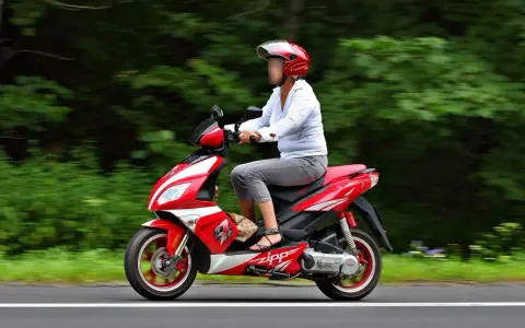 Kobieta w kasku jedzie czerwonym skuterem po autostradzie, otoczona zielenią.