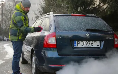 Mężczyzna w zimowej kurtce czyści zamarzniętą szybę auta. Dym z rury wydechowej sugeruje, że to diesel i właśnie próbuje się dowiedzieć, jak odpalić diesla w mroźny dzień.