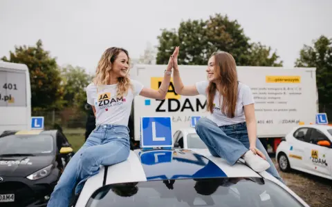 Dwie uśmiechnięte dziewczyny w koszulkach "JA ZDAM SZKOŁA JAZDY" dają sobie piątkę, siedząc na dachu samochodu szkoleniowego.