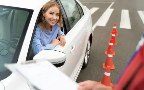 Uśmiechnięta kobieta w samochodzie czeka na rozpoczęcie egzaminu wewnętrznego na prawo jazdy. Przed nią stoją pomarańczowe pachołki.