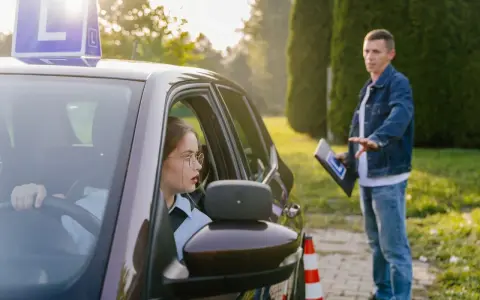 Młoda kobieta w okularach za kierownicą, z literą "L" na dachu, uczy się jeździć. Instruktor z tablicą daje wskazówki. To jej kontynuacja prawa jazdy po latach.