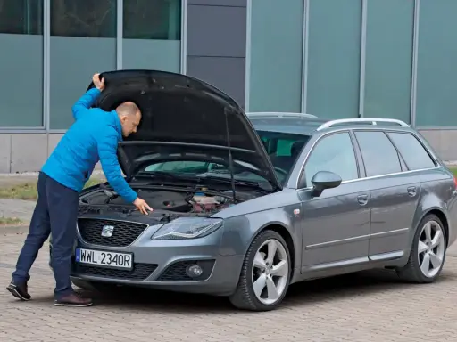 Mężczyzna w niebieskiej kurtce sprawdza silnik szarego kombi. To ważny moment przed kupnem samochodu, by upewnić się, że wszystko działa.