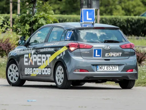 Szara Skoda i20 z oznaczeniami "Szkoła Jazdy EXPERT" i literą "L" na dachu i bokach. To auto do nauki jazdy, idealne dla przyszłego kierowcy.