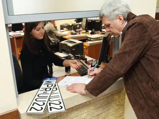 Mężczyzna podpisuje wniosek o rejestrację pojazdu, urzędniczka podaje mu dokumenty. Obok leżą nowe tablice rejestracyjne.
