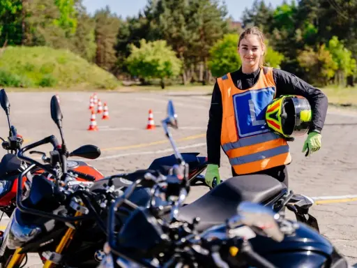Młoda kobieta w kamizelce z literą "L" i kaskiem w ręku, gotowa do jazdy na motocyklu na placu manewrowym. To jej pierwsze kroki na drodze, a wkrótce będzie mogła jeździć na A1.