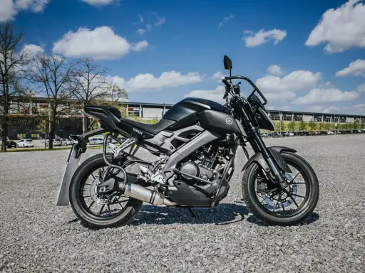 Czarny motocykl Yamaha MT-125, model kat a1, zaparkowany na żwirowym placu przed budynkiem.