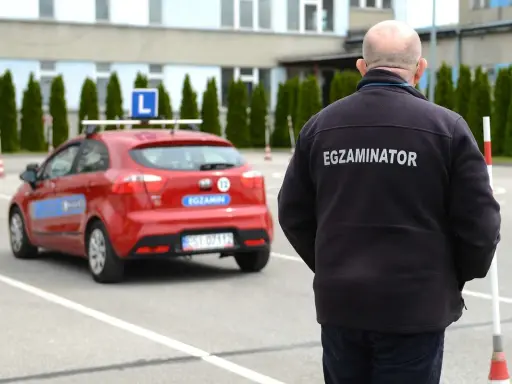 Egzaminator w czarnej kurtce z napisem "EGZAMINATOR" obserwuje czerwony samochód z literą "L" na dachu. Jak odwołać egzamin na prawo jazdy infocar?