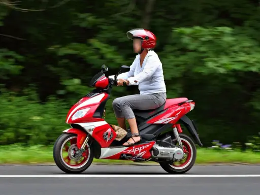 Kobieta w kasku jedzie czerwonym skuterem po autostradzie, otoczona zielenią.