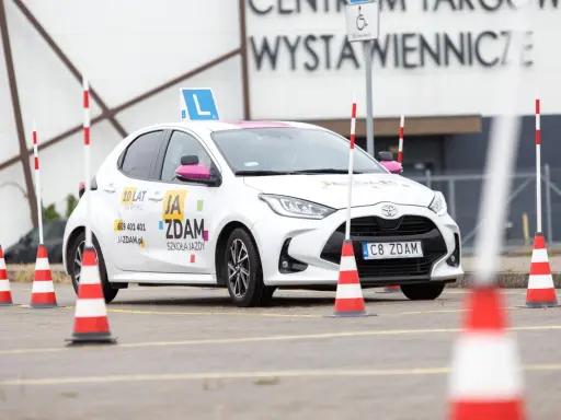 Biały samochód szkoleniowy z literą "L" na dachu i pachołkami na placu manewrowym. To miejsce, gdzie nauczysz się, jak wypełnić pkk.