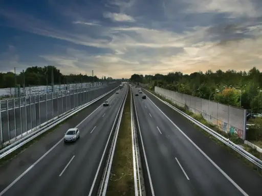 Szeroka autostrada z samochodami, wjazd na autostradę w tle, po bokach ekrany akustyczne i drzewa.