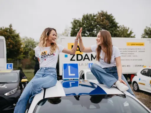 Dwie uśmiechnięte dziewczyny w koszulkach "JA ZDAM SZKOŁA JAZDY" dają sobie piątkę, siedząc na dachu samochodu szkoleniowego.