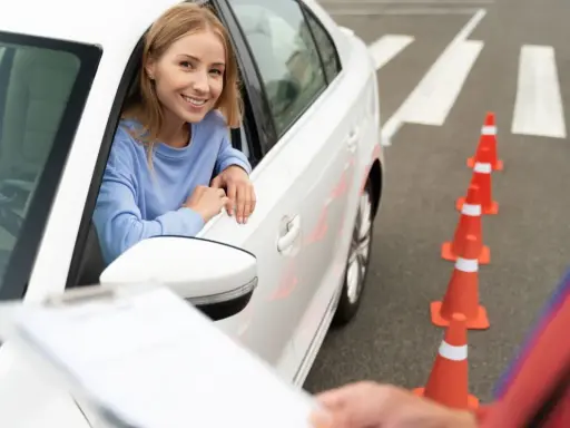 Uśmiechnięta kobieta w samochodzie czeka na rozpoczęcie egzaminu wewnętrznego na prawo jazdy. Przed nią stoją pomarańczowe pachołki.