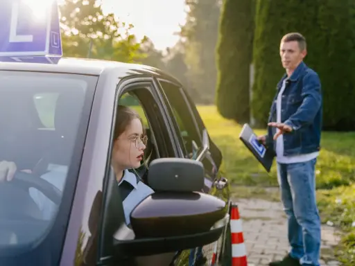 Młoda kobieta w okularach za kierownicą, z literą "L" na dachu, uczy się jeździć. Instruktor z tablicą daje wskazówki. To jej kontynuacja prawa jazdy po latach.