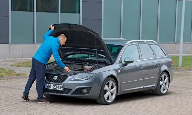 Mężczyzna w niebieskiej kurtce sprawdza silnik szarego kombi. To ważny moment przed kupnem samochodu, by upewnić się, że wszystko działa.