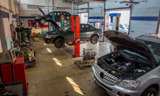Warsztat samochodowy z kilkoma autami na podnośnikach. Widać m.in. ciemny sedan, który wygląda jak p1351 peugeot, oraz srebrny SUV z otwartą maską.