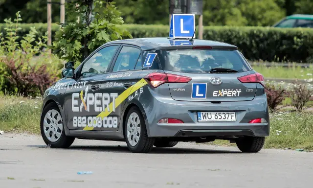 Szara Skoda i20 z oznaczeniami "Szkoła Jazdy EXPERT" i literą "L" na dachu i bokach. To auto do nauki jazdy, idealne dla przyszłego kierowcy.