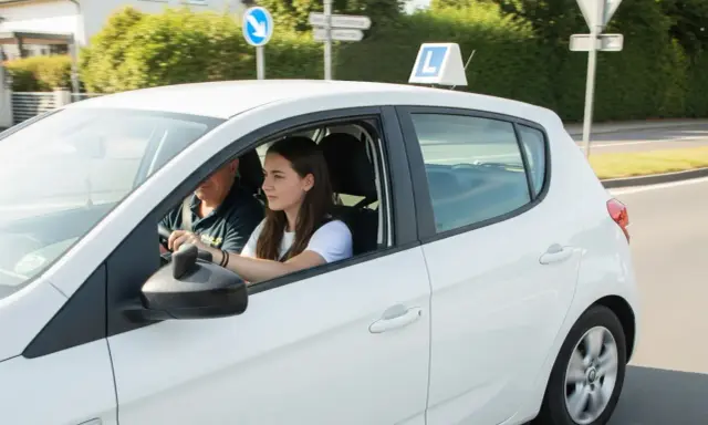 Młoda dziewczyna uczy się jeździć, a obok siedzi instruktor. Znak "L" na dachu sugeruje, że to nauka jazdy. Od kiedy można robić prawo jazdy?