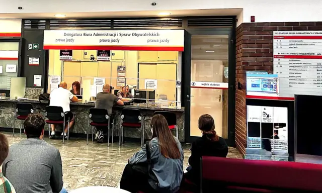 W urzędzie ludzie czekają na obsługę. Na tablicy informacja o tym, jak sprawdzić swój numer PKK online, dotyczy spraw związanych z prawem jazdy.