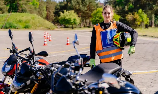 Młoda kobieta w kamizelce z literą "L" i kaskiem w ręku, gotowa do jazdy na motocyklu na placu manewrowym. To jej pierwsze kroki na drodze, a wkrótce będzie mogła jeździć na A1.
