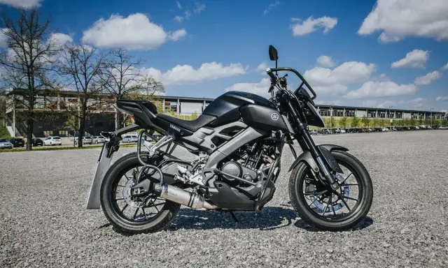 Czarny motocykl Yamaha MT-125, model kat a1, zaparkowany na żwirowym placu przed budynkiem.