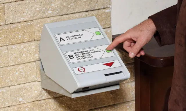 Dłoń naciska przycisk na automacie do wydawania dowodów rejestracyjnych, ułatwiając wymianę dowodu rejestracyjnego online.
