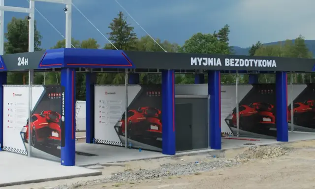Nowoczesna myjnia bezdotykowa z niebieskimi elementami i reklamami czerwonych samochodów.