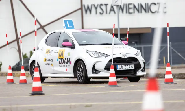 Biały samochód szkoleniowy z literą "L" na dachu i pachołkami na placu manewrowym. To miejsce, gdzie nauczysz się, jak wypełnić pkk.